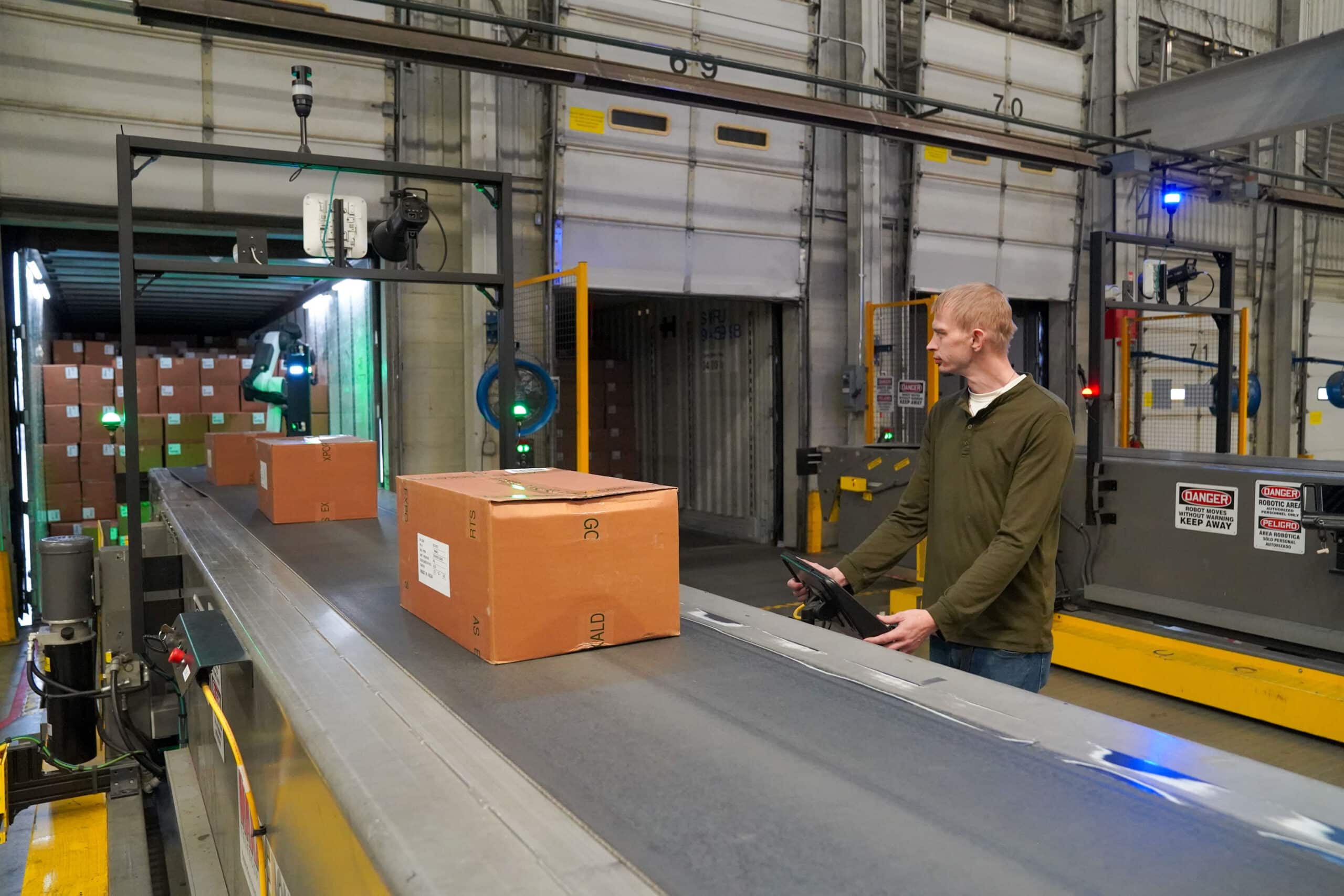 Worker in front of conveyor belt watches robot unloading boxes.